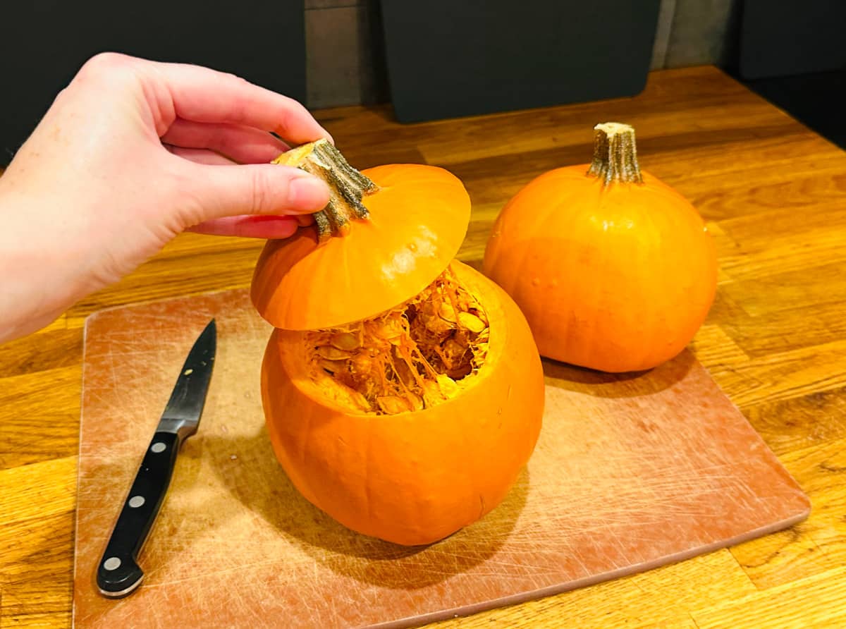 Lid being removed from a small pumpkin sitting between a knife and another pumpkin.