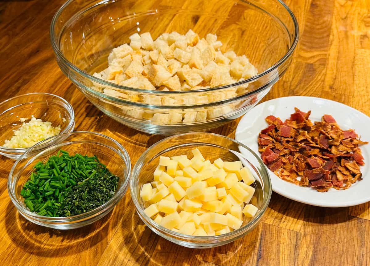 Five separate glass bowls containing bread cubes, minced garlic, chopped chives and thyme, cheese cubes, and bacon bits.