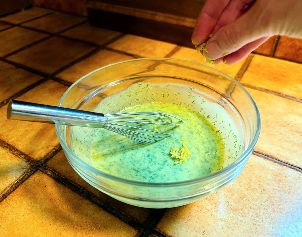 Lemon zest being sprinkled into a glass bowl containing yellow sauce flecked with dill and a metal whisk.