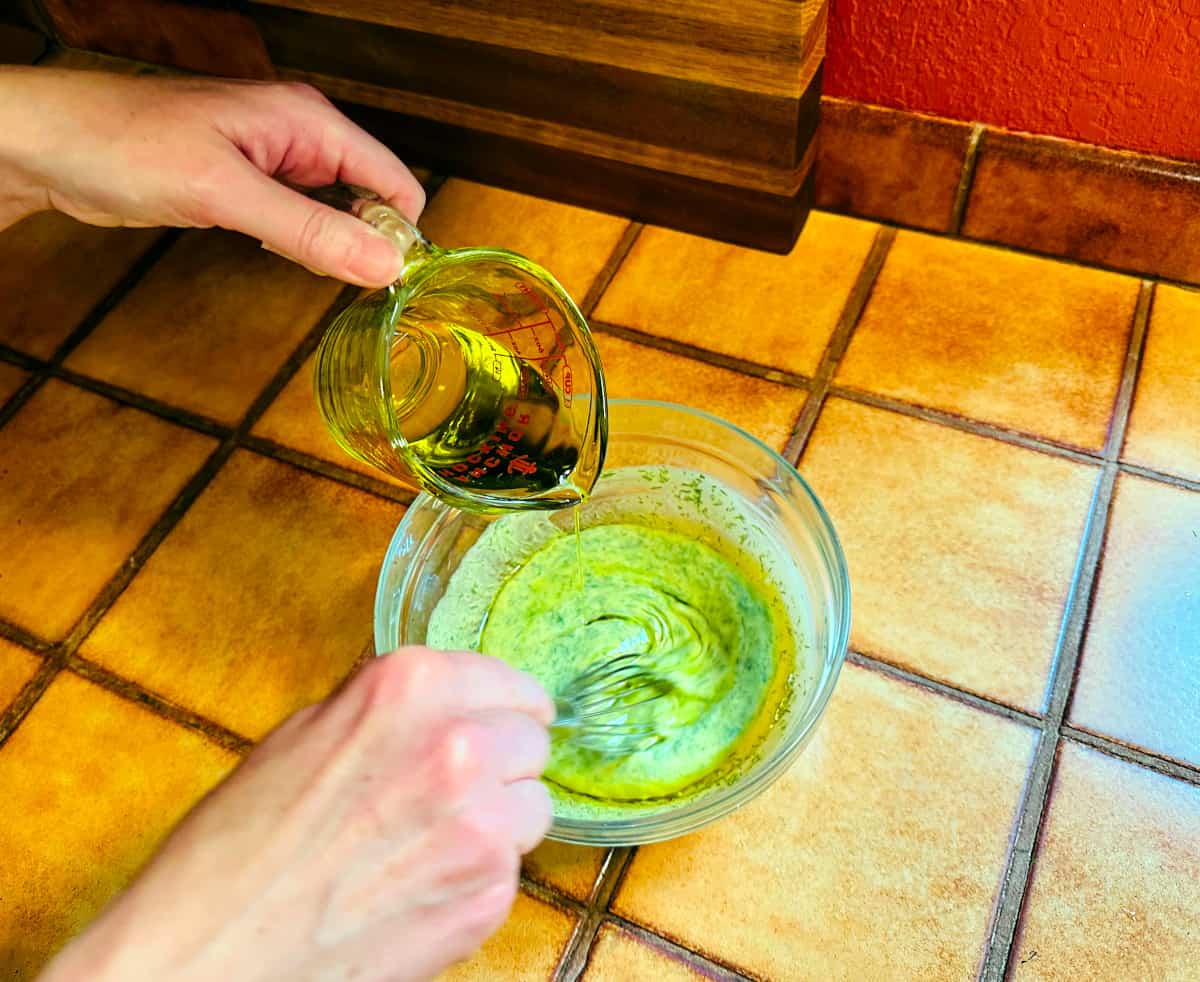 Oil being poured from a glass measuring cup into a glass bowl containing yellow sauce flecked with green being stirred with a whisk.