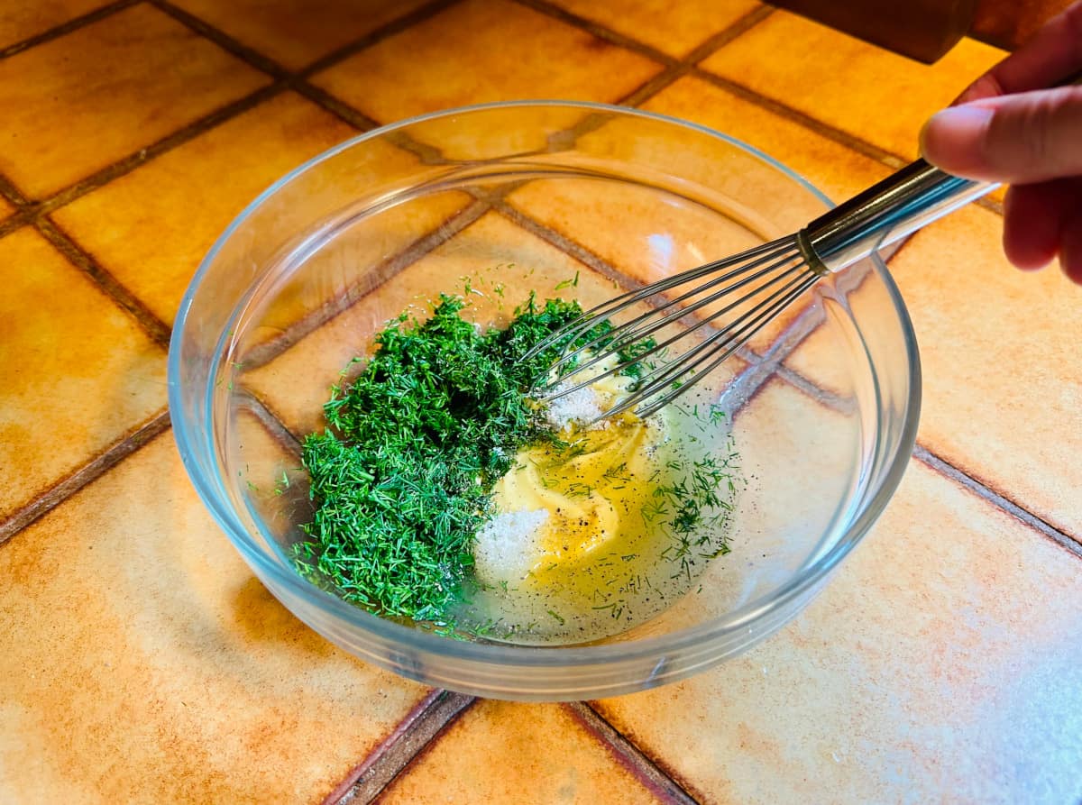 Chopped dill, mustard, lemon juice and zest, and sugar being stirred in a glass bowl with a metal whisk.
