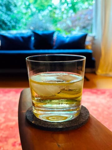 White Negroni in an old fashioned glass with a lemon twist sitting on the wooden arm rest of a chair in a living room with a big picture window, navy chaise lounge covered in pillows, and red carpet.