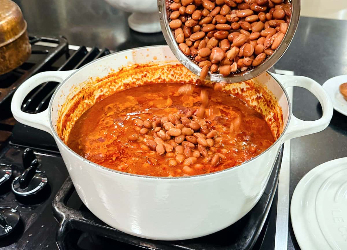 Pinto beans being tipped from a small steel colander into chili simmering in a large white Dutch oven.
