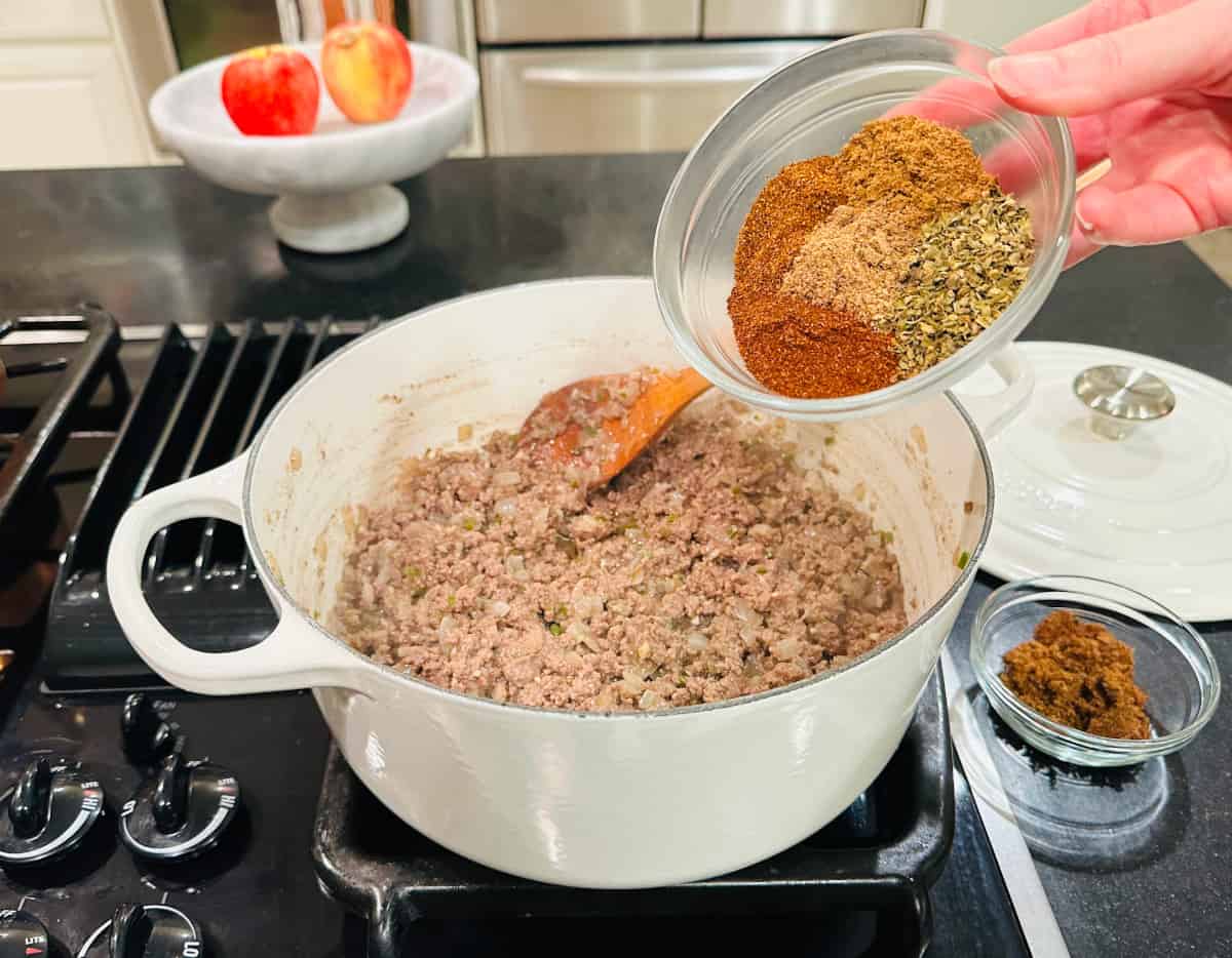 Chili powders, oregano, cumin, and coriander in a small glass bowl being tipped into browned beef and onion mixture in a large white Dutch oven.