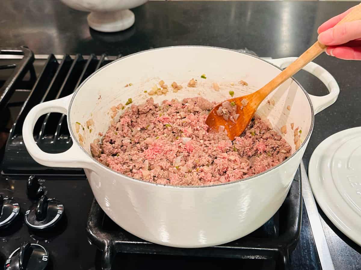 Ground beef browning in a large white Dutch oven while being stirred with a wooden spoon.