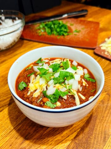 Chili con carne topped with shredded cheddar, chopped white onion, and chopped cilantro in a white bowl with a navy stripe around the rim.