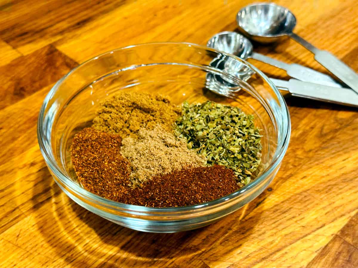 Chili powders, cumin, oregano, and coriander in a small glass bowl next to a set of steel measuring spoons.