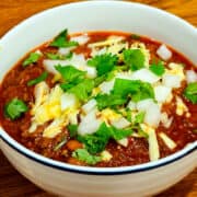 Chili con carne topped with shredded cheddar, chopped white onion, and chopped cilantro in a white bowl with a navy stripe around the rim.