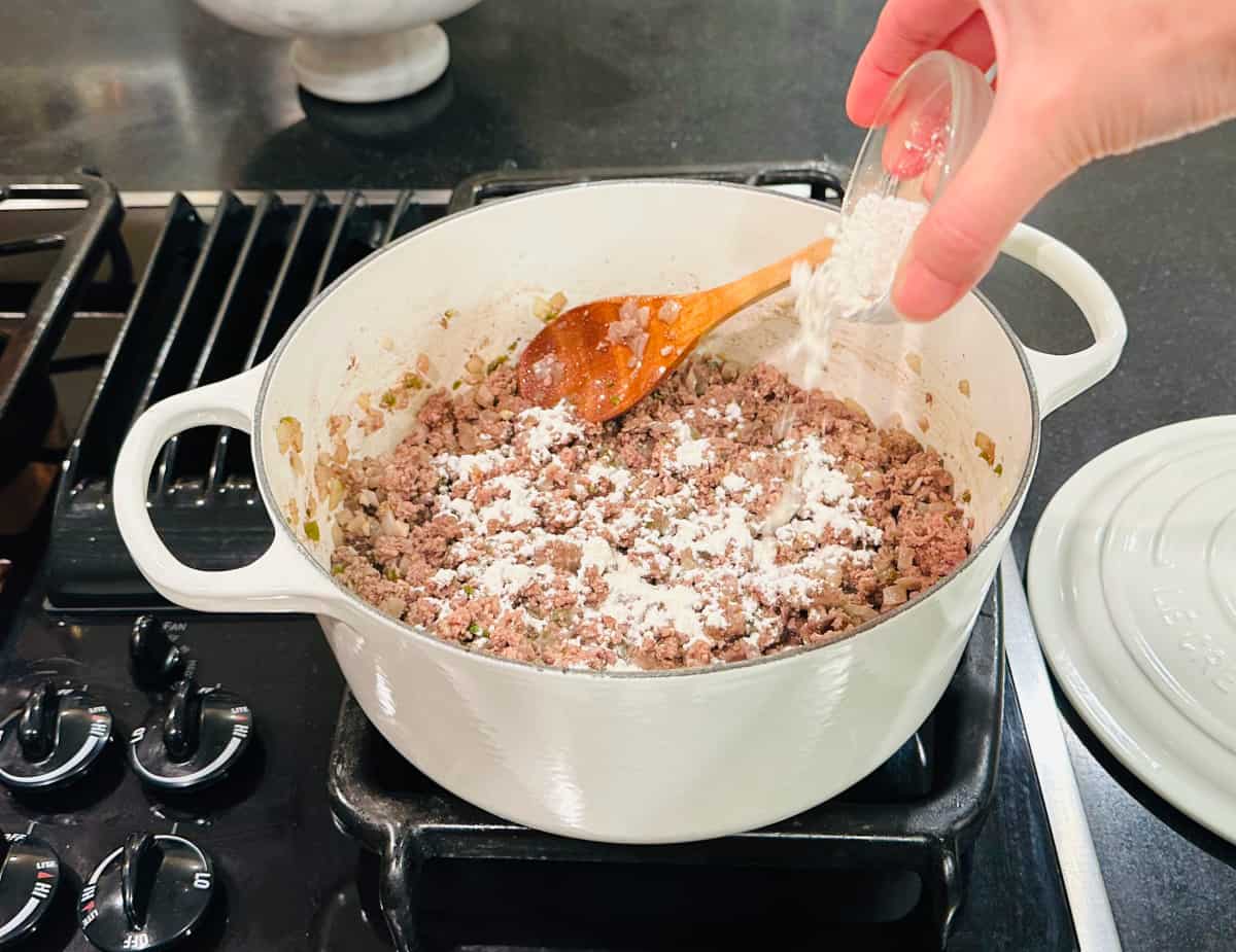 Flour being sprinkled from a small glass bowl into browned ground beef in a large white Dutch oven.