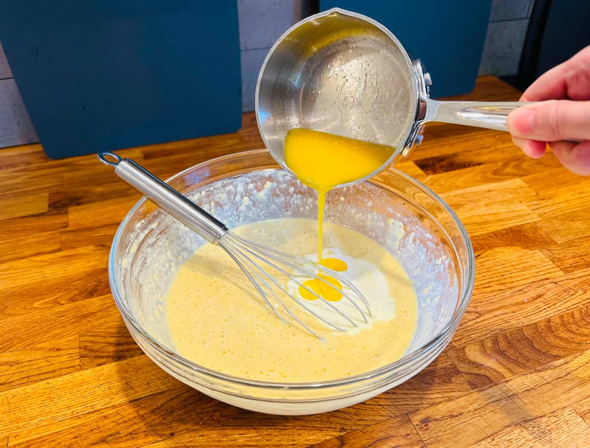Melted butter being poured form a small steel saucepan into yellow batter in a glass bowl with a metal whisk.