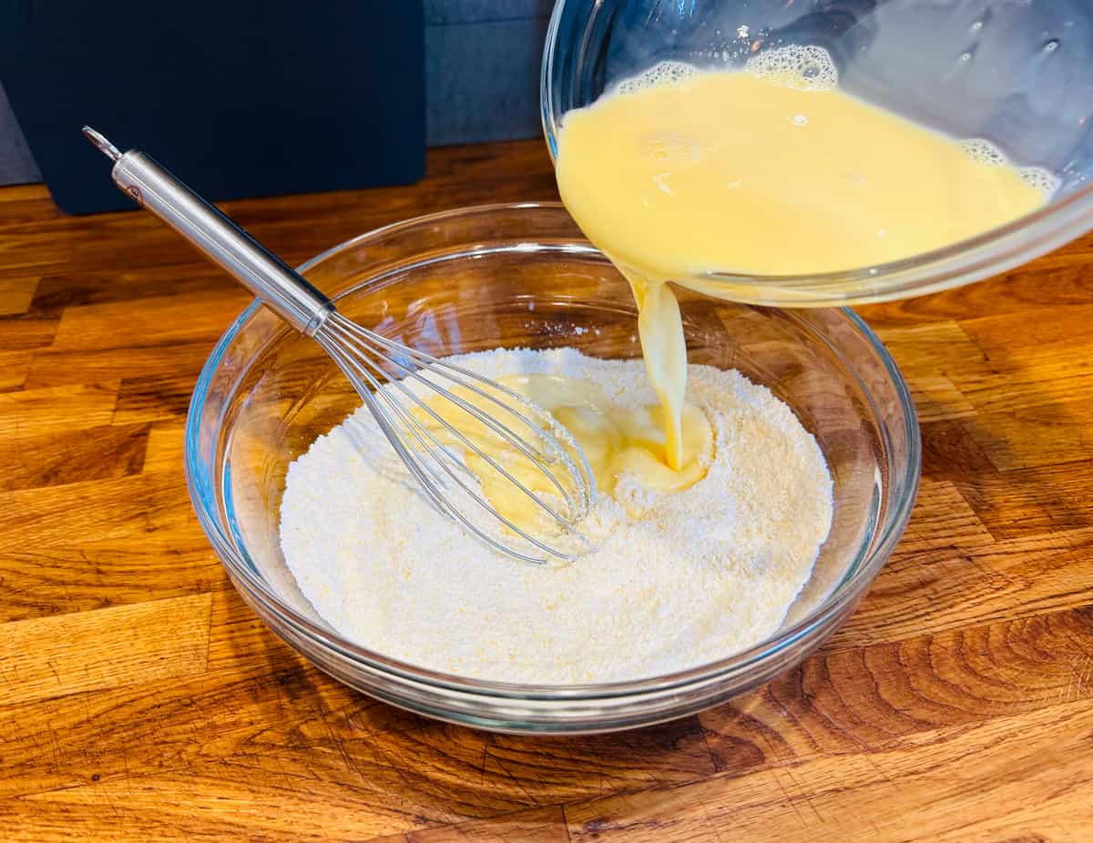 Yellow liquid being poured out of a glass bowl into a larger glass bowl containing dry ingredient mixture and a metal whisk.