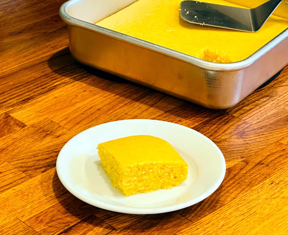 Slice of yellow corn bread on a white plate in front of a metal pan of cornbread with a small metal spatula resting inside.