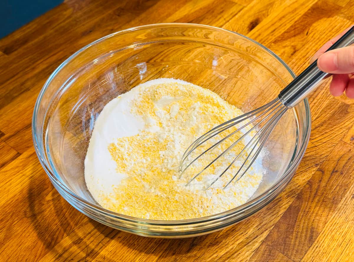 Cornmeal, flour, salt, sugar, and baking powder being combined in a glass bowl with a metal whisk.