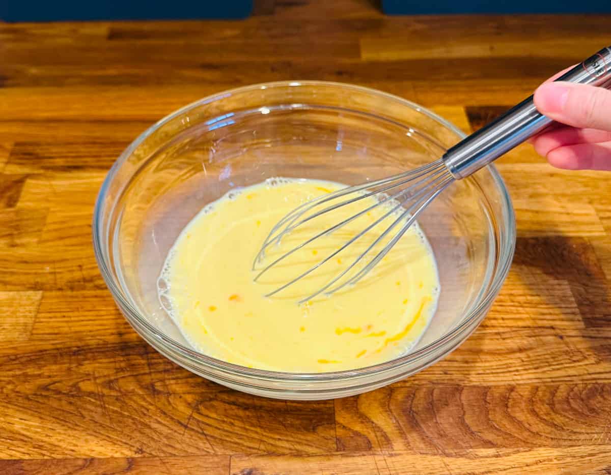 Yellow mixture of eggs and milk being combined with a metal whisk in a glass bowl.