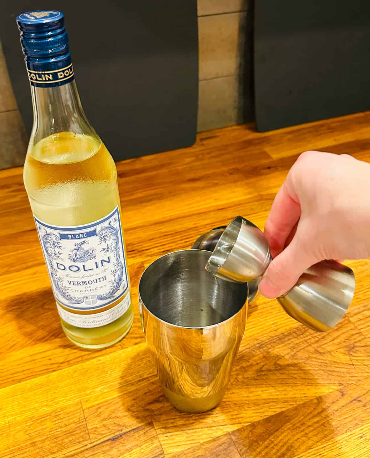 Clear liquid being poured from a steel measuring jigger into a cocktail shaker next to a bottle of Dolin blanc vermouth.
