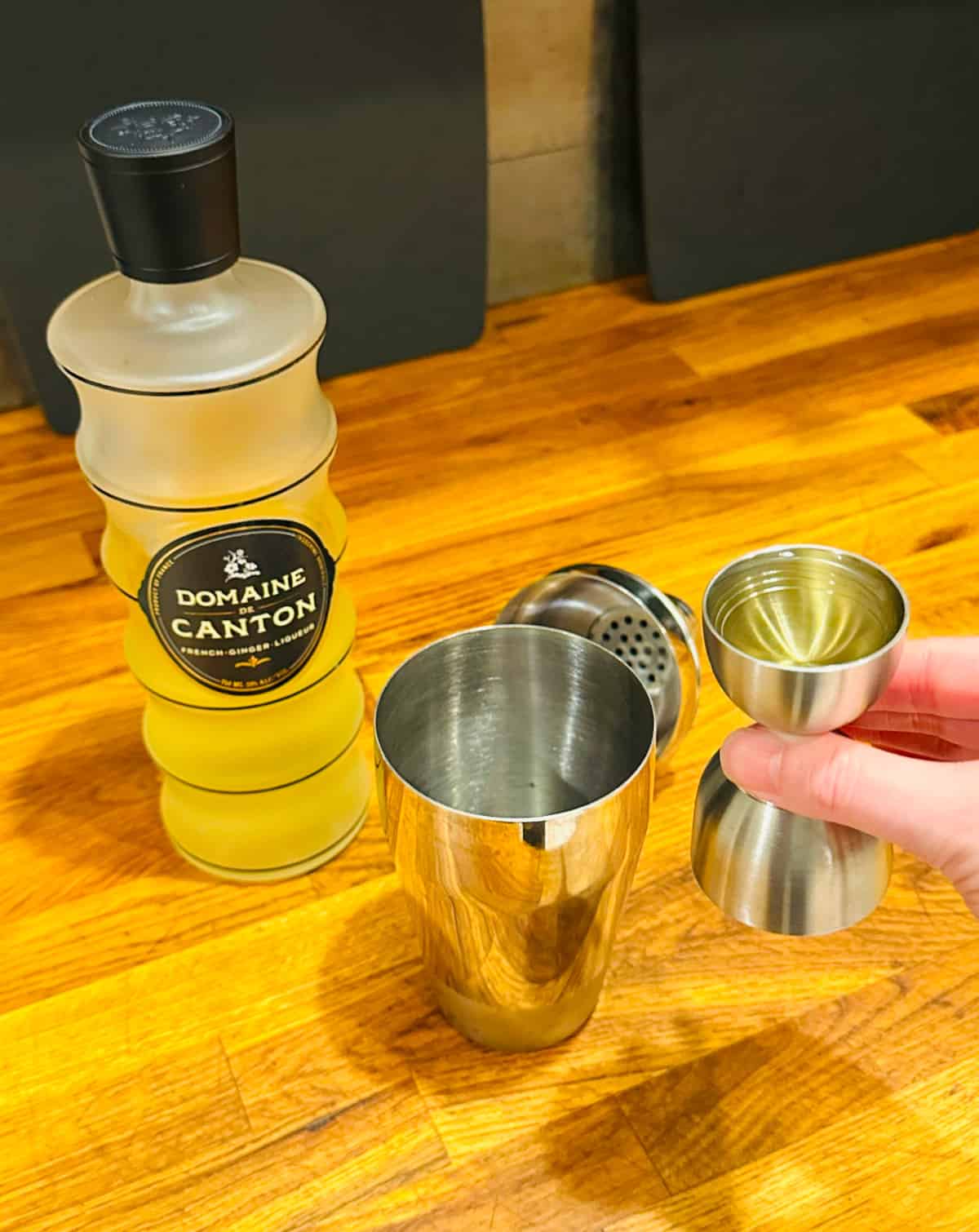 Pale yellow liquid in a steel measuring jigger next to a cocktail shaker and a bottle of Domaine de Canton ginger liqueur.