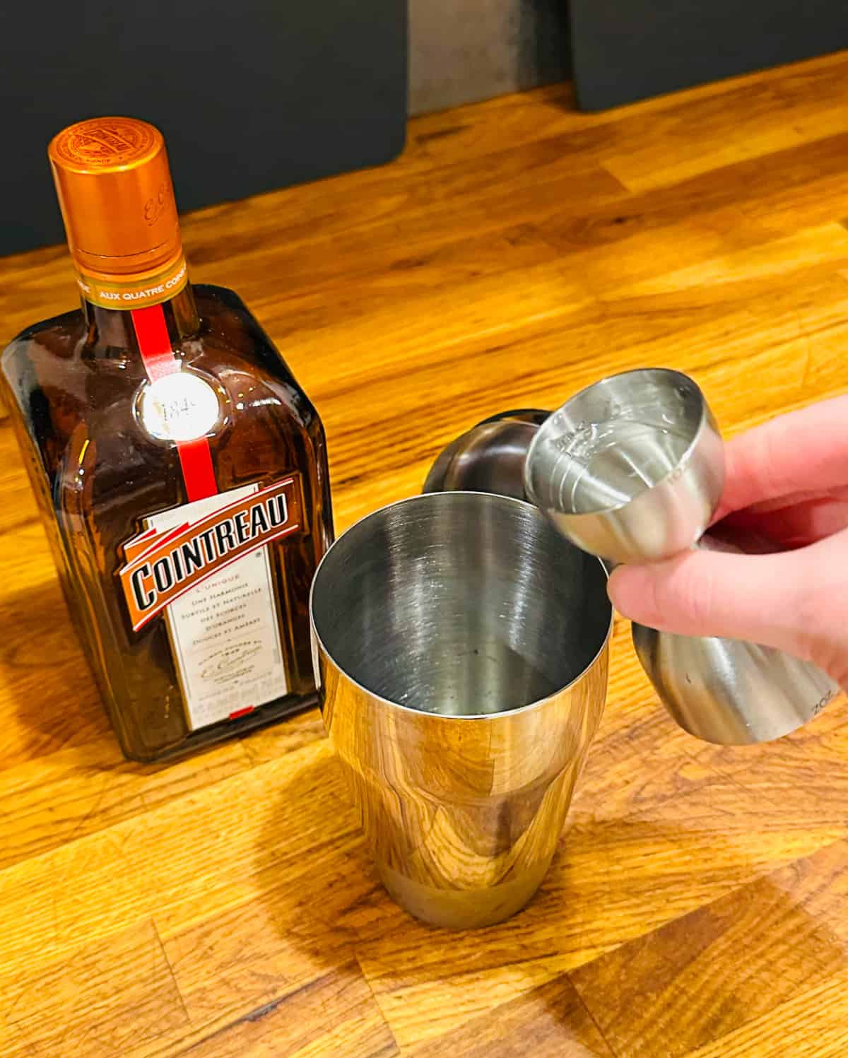Clear liquid being poured from a steel measuring jigger into a cocktail shaker next to a bottle of Cointreau.