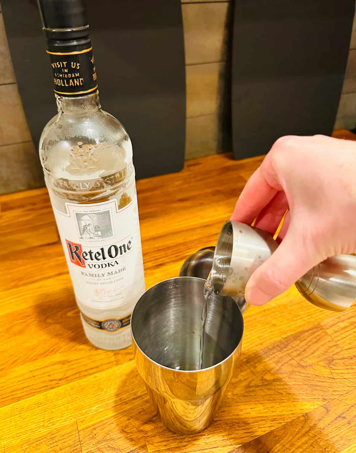 Clear liquid being poured from a steel measuring jigger into a cocktail shaker next to a bottle of Ketel One vodka.