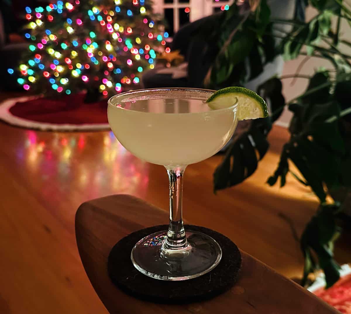 Ginger martini garnished with a lime wedge and sitting on a wooden arm rest in front of a Christmas tree.