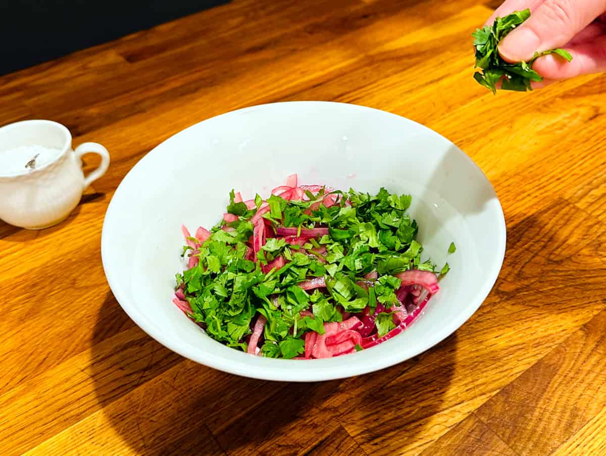 Chopped cilantro being sprinkled over steeped and drained red onions in a white bowl next to a small cup of sea salt.