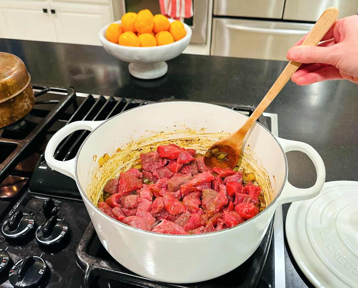 Lamb being stirred with a wooden spoon into onions in a large white pot on the stove.