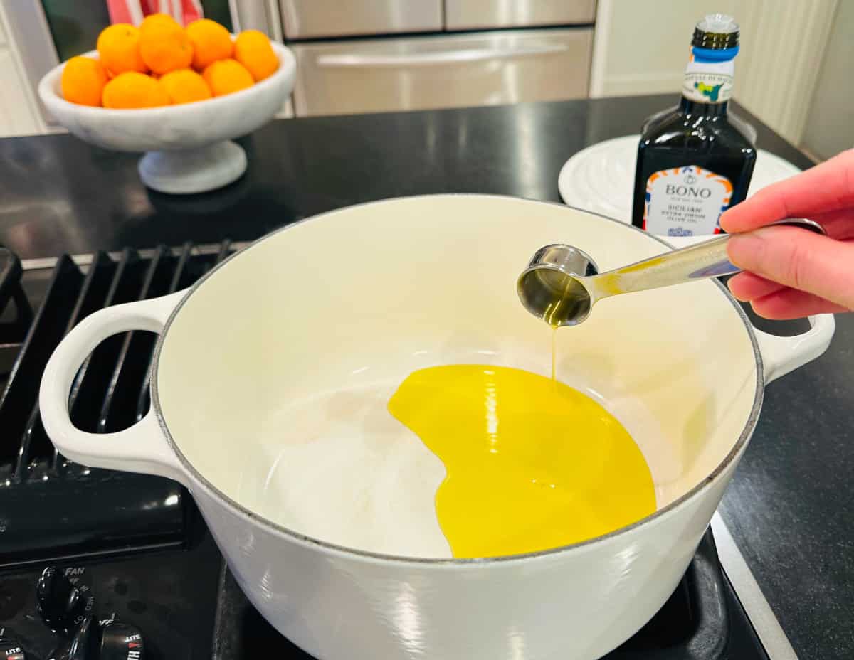 Olive oil being tipped from a steel measuring spoon into a large white pot on the stove.