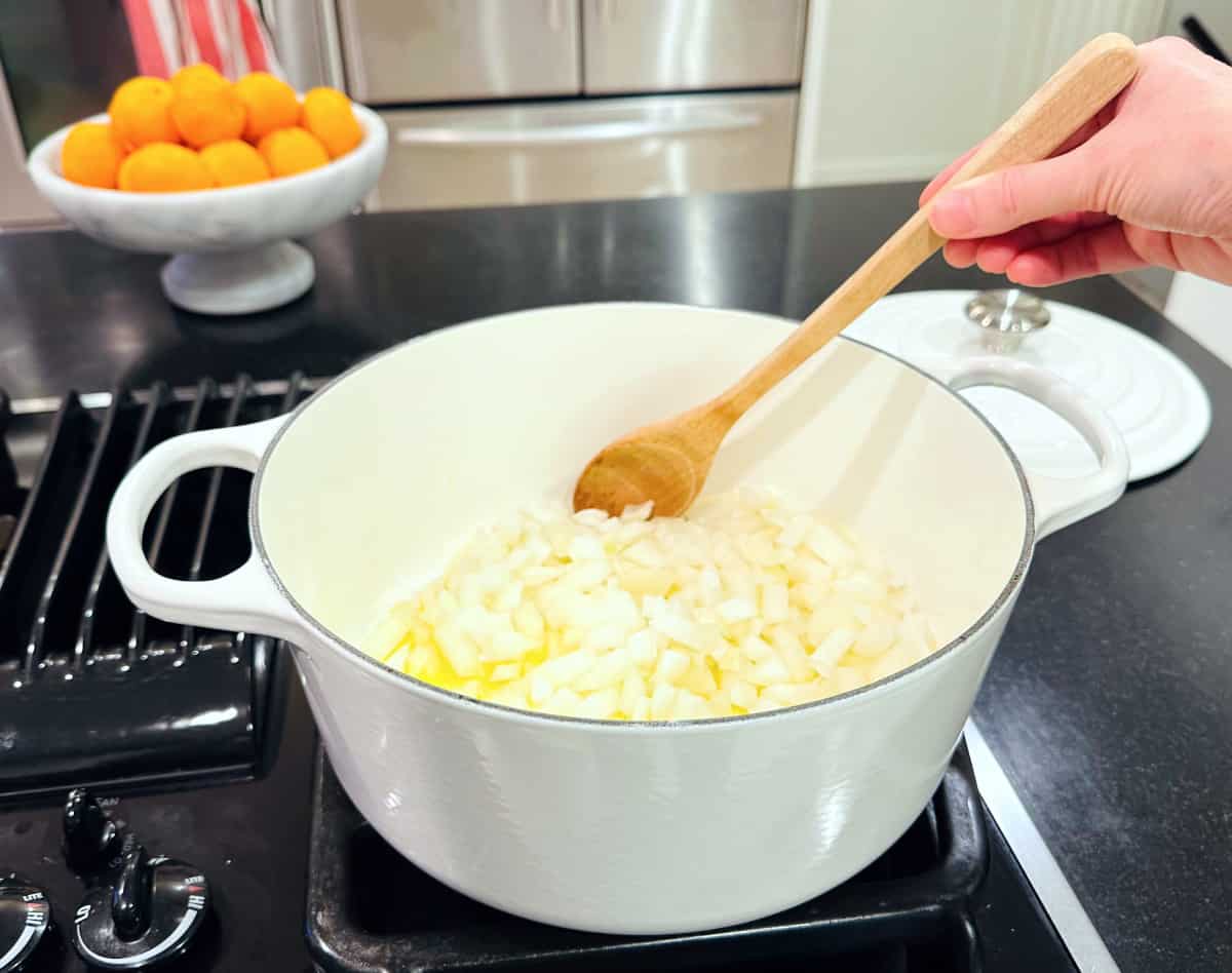 Onions being stirred with a wooden spoon in a large white pot on the stove.
