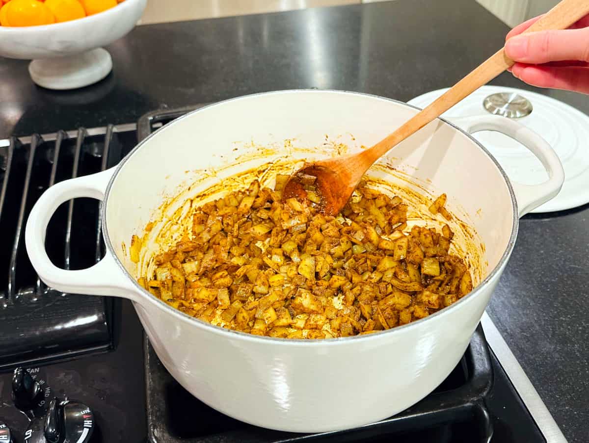 Spices being stirred with a wooden spoon into onions in a large white pot on the stove.