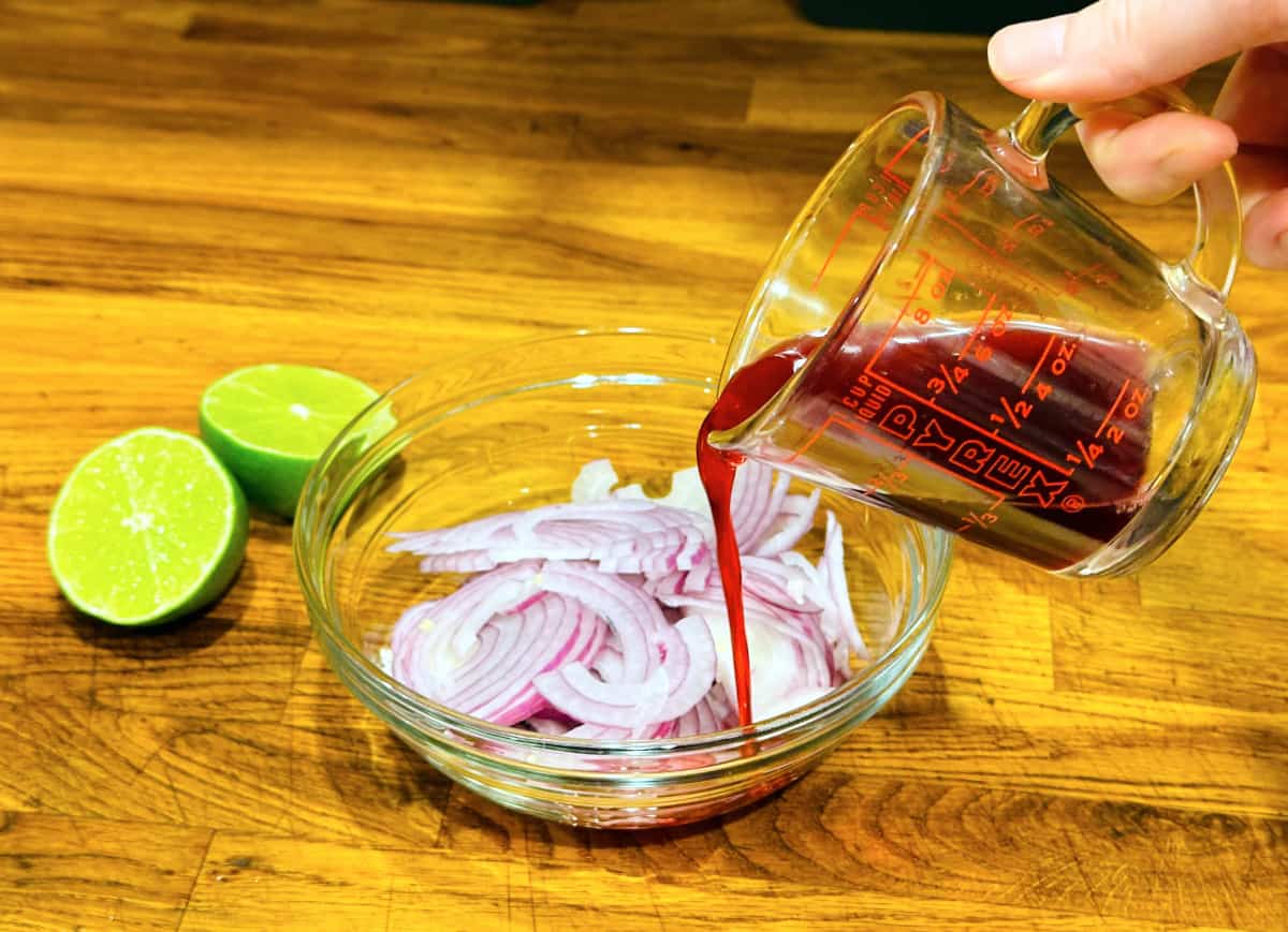 Pomegranate juice being poured from a glass measuring cup over sliced red onions in a glass bowl next to a halved lime.