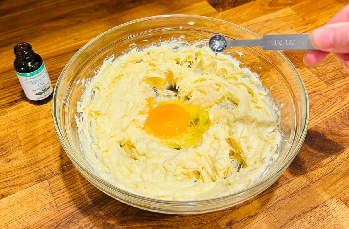 Clear liquid being tipped from a steel measuring spoon into a glass bowl containing a fluffy yellow mixture and egg.