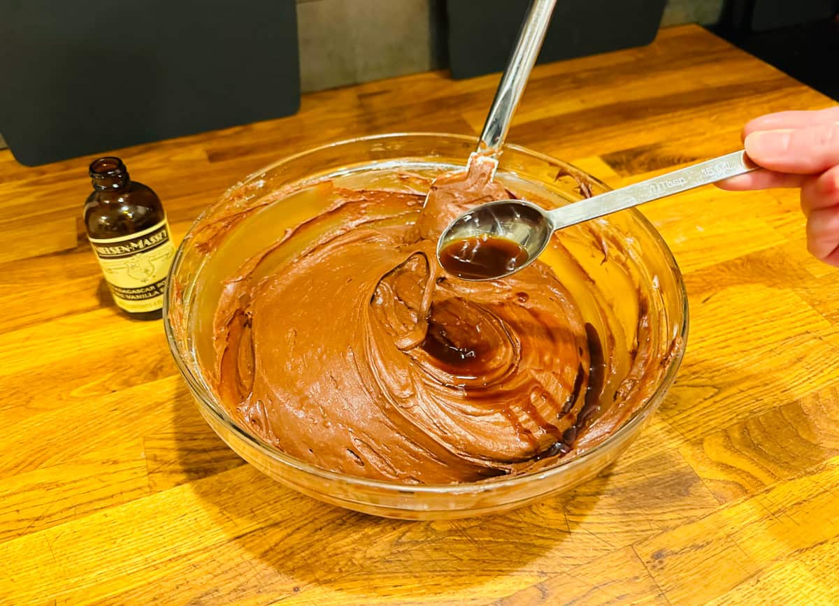 Vanilla extract being tipped from a steel measuring spoon into chocolate batter in a glass bowl.