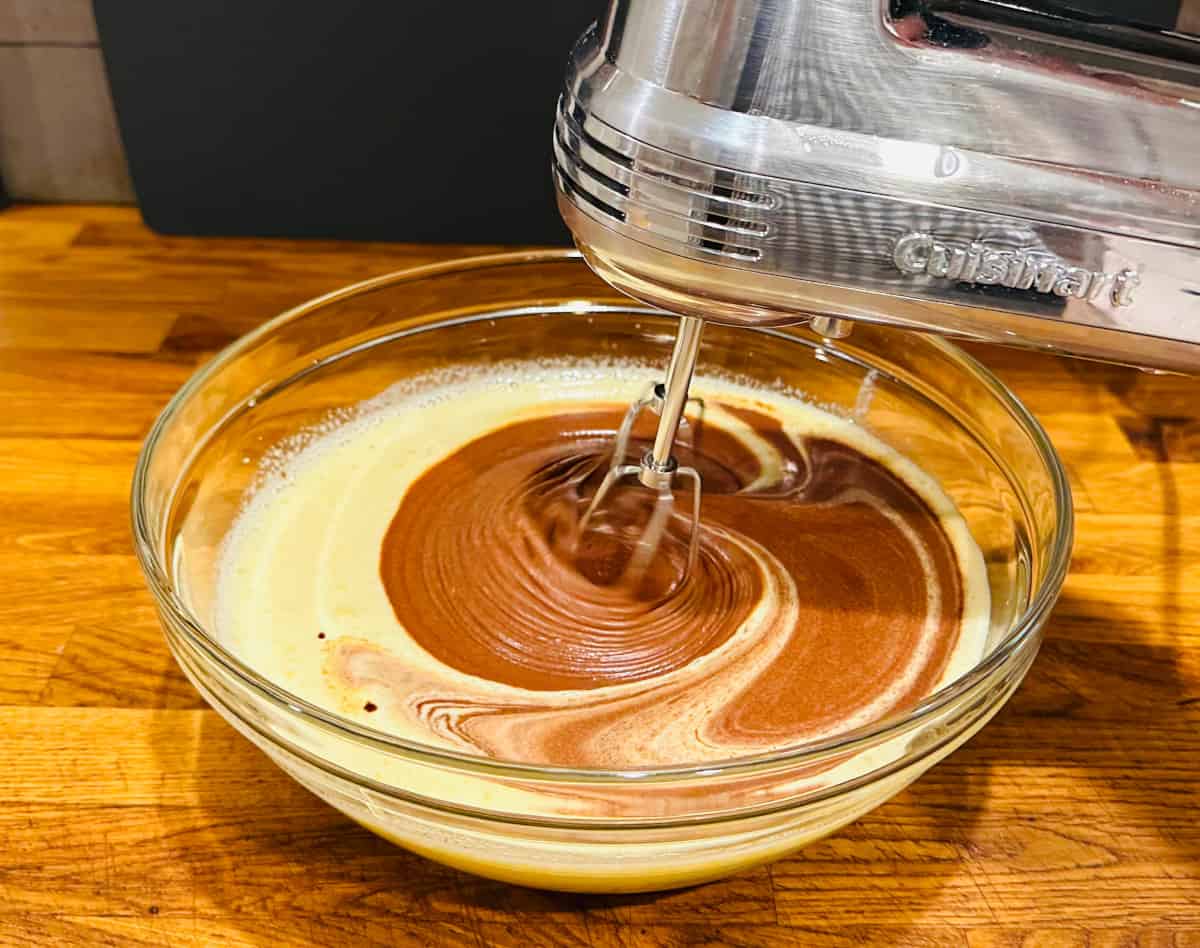 Chocolate being mixed into sugar and egg mixture with an electric hand mixer in a large glass bowl.