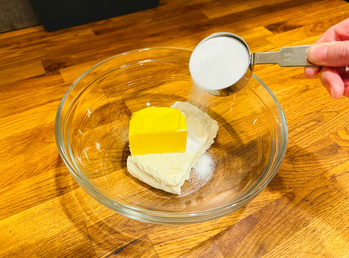 Sugar being poured from a steel measuring cup into a glass bowl containing cream cheese and butter.