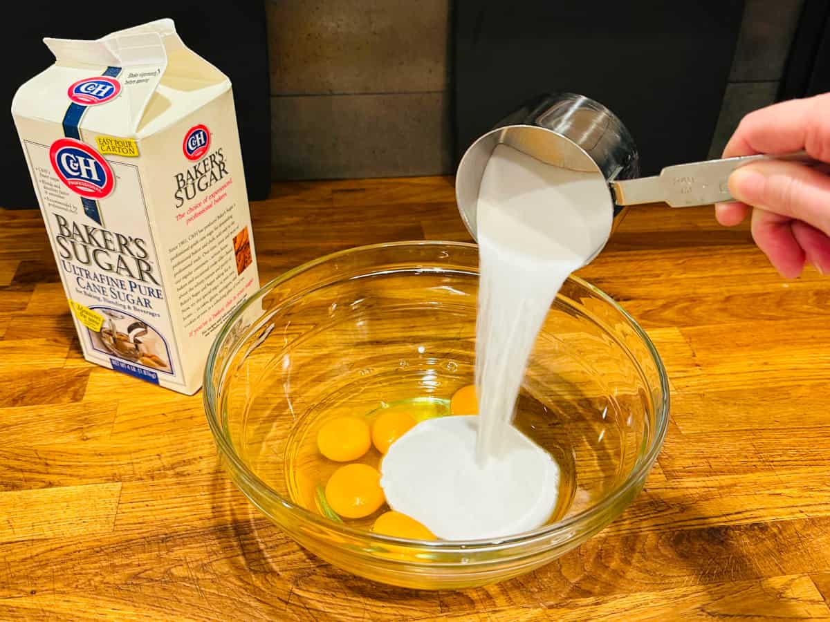 Sugar being poured from steel measuring cup into eggs in a glass bowl.