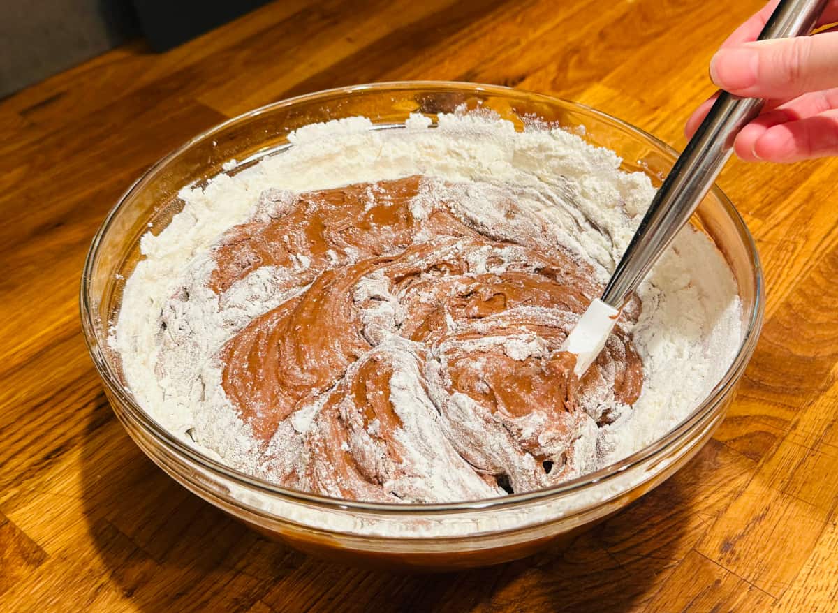 Flour mixture being stirred into chocolate batter with a silicone spatula in a large glass bowl.