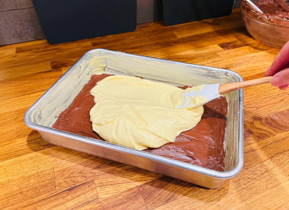 Cream cheese filling being spread over chocolate batter in a greased metal baking pan.