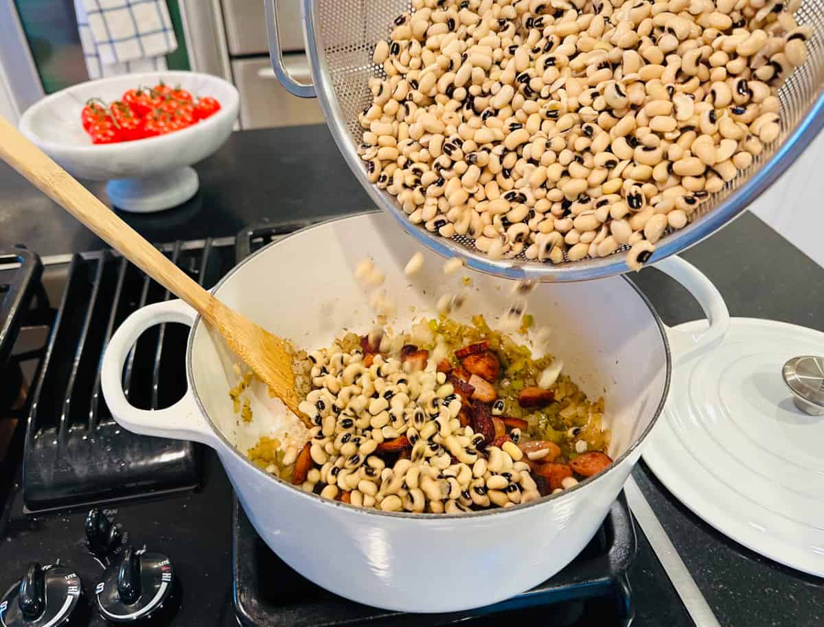 Black eyed peas being tipped from a metal colander into a large white pot containing cooked vegetables and browned sausage.