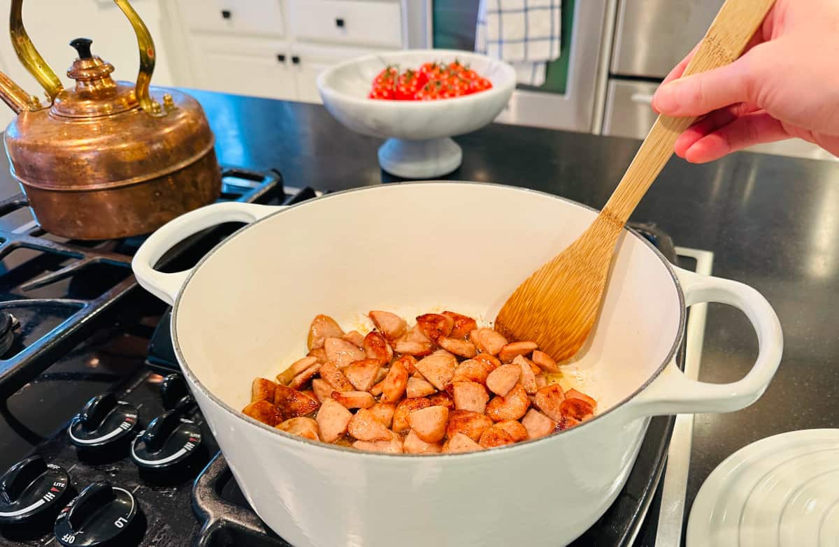 Sausage being stirred with a wooden spoon and browned in a large white pot.