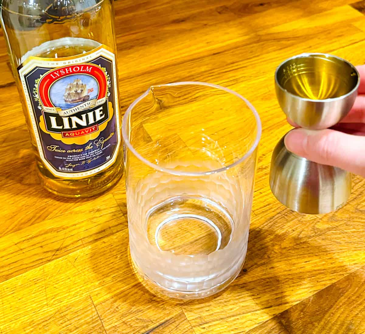 Pale golden liquid in a steel measuring jigger next to a mixing glass and a bottle of Linie aquavit.