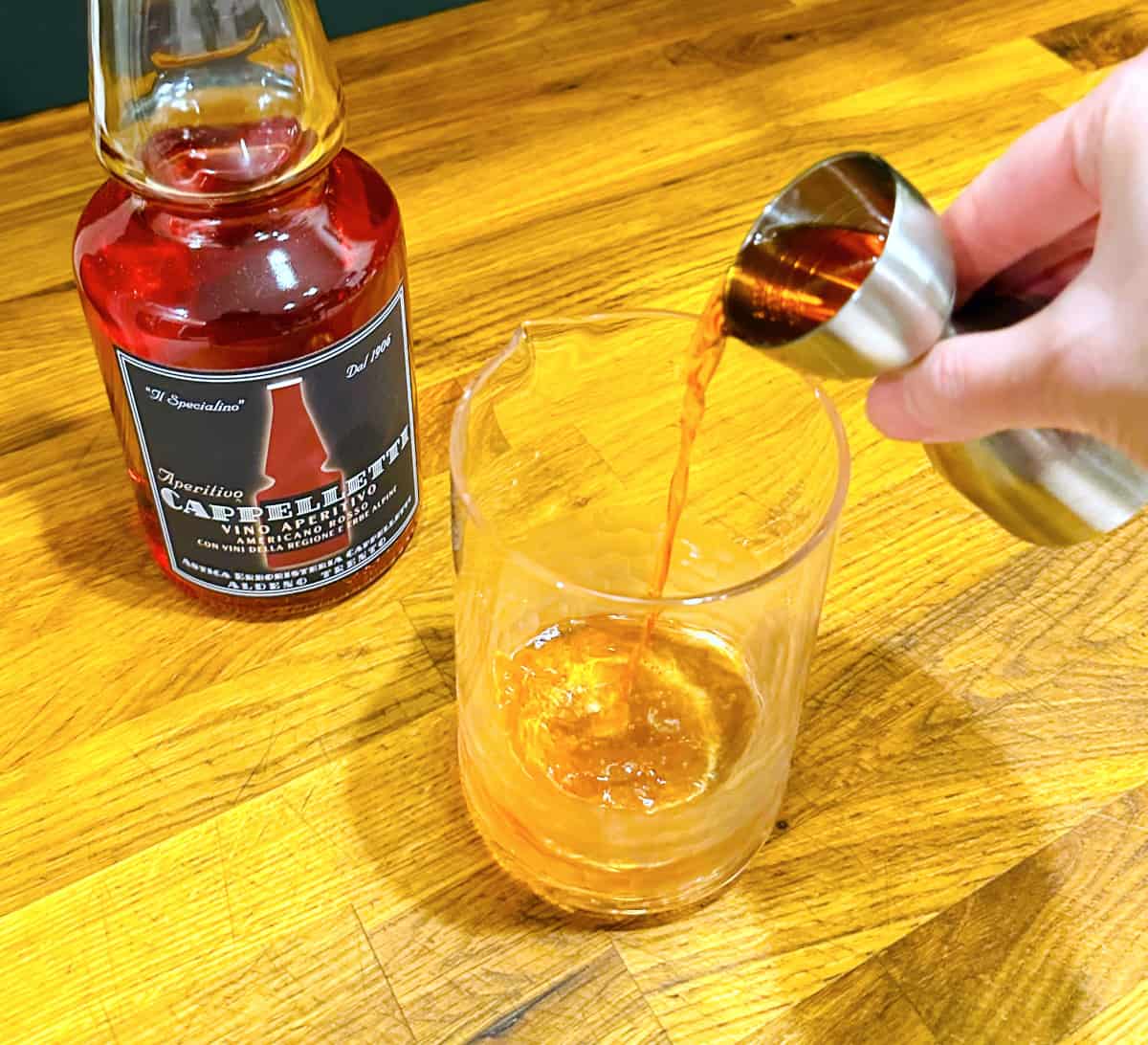 Pale red liquid being poured from a steel measuring jigger into a mixing glass next to a bottle of Cappelletti.