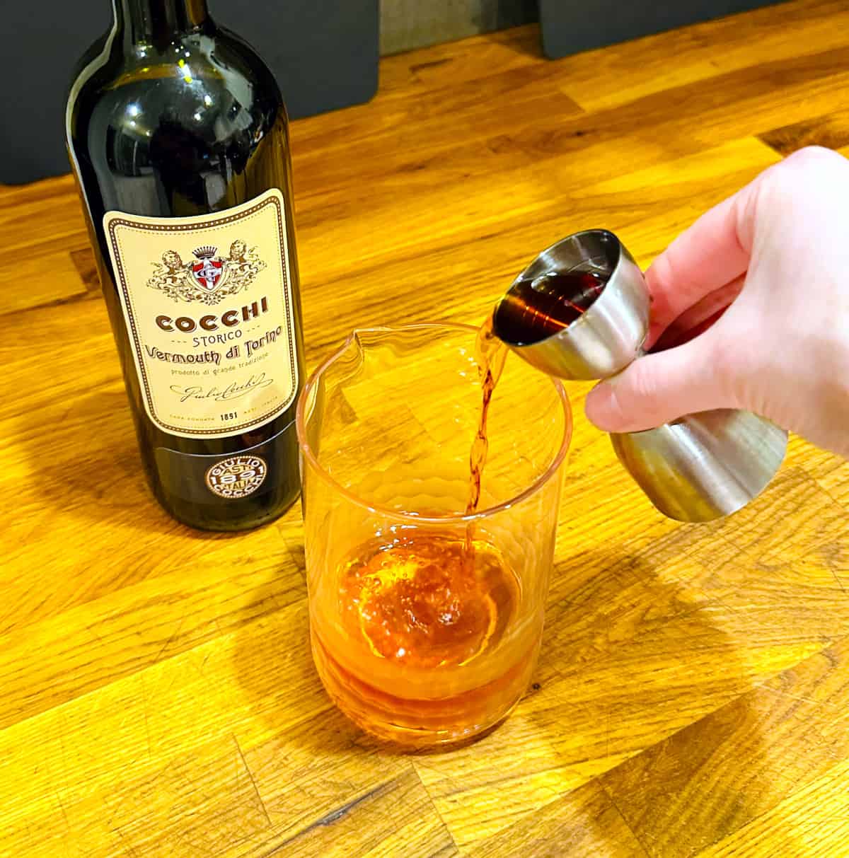 Pale brown liquid being poured from a steel measuring jigger into a mixing glass next to a bottle of Cocchi Vermouth di Torino.