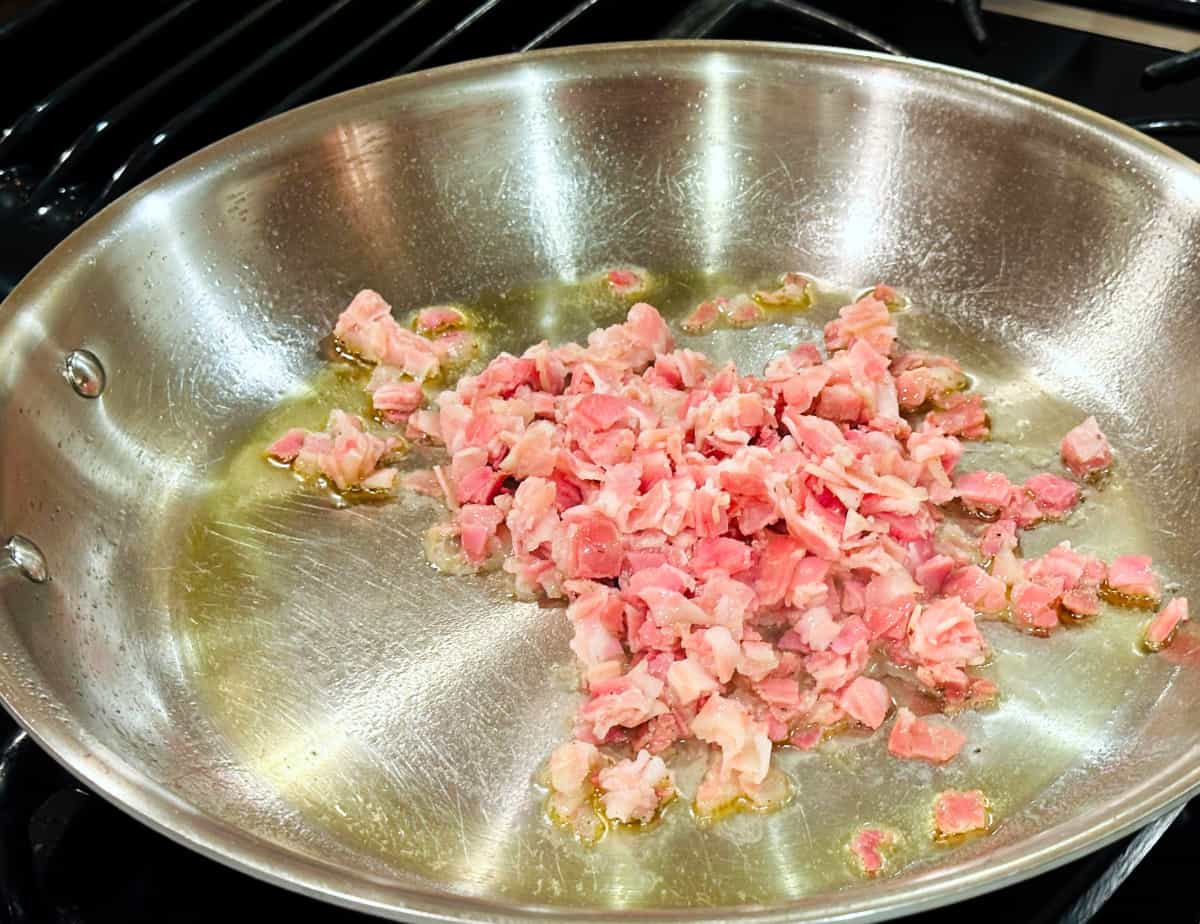 Chopped pancetta cooking in a large steel skillet coated with olive oil.