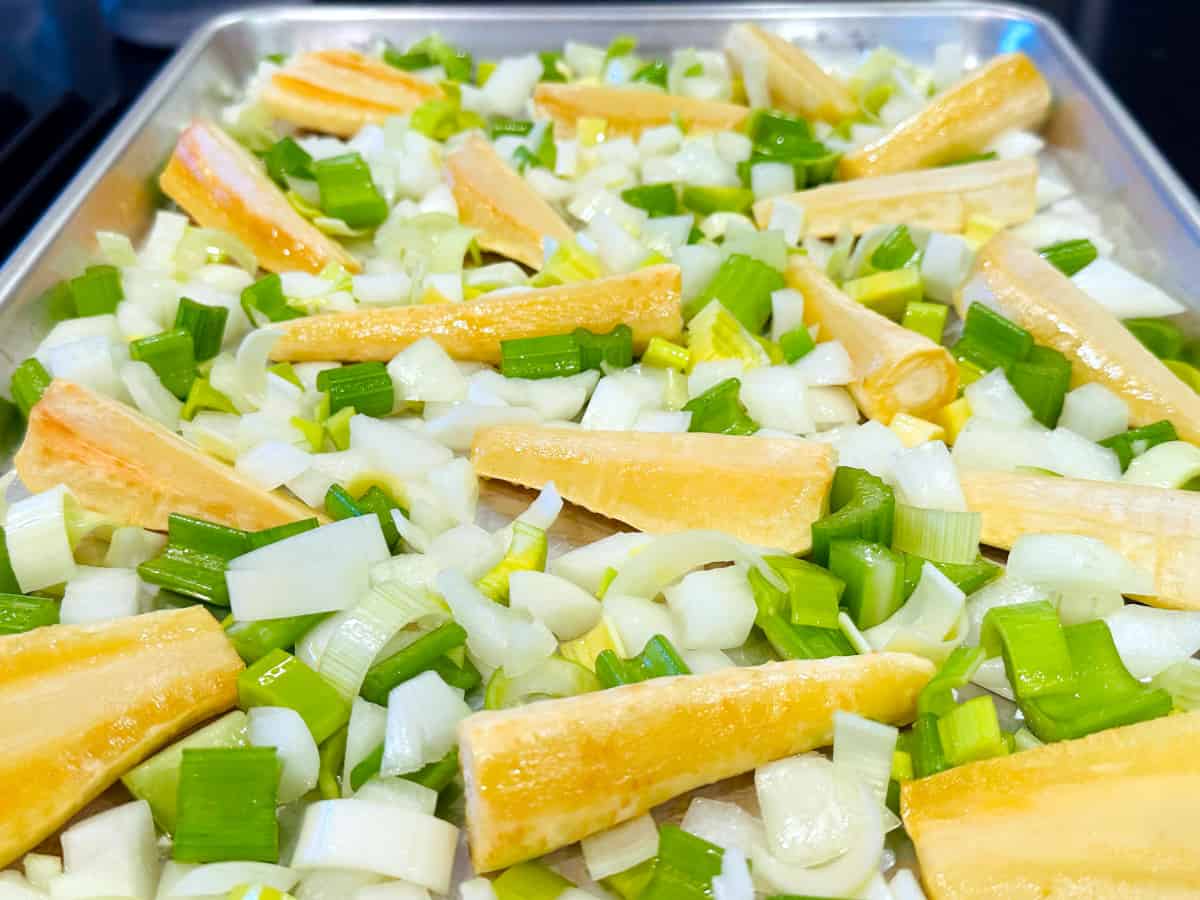 Partially roasted parsnips with chopped onion, celery, and leek scattered around them on a metal baking sheet.