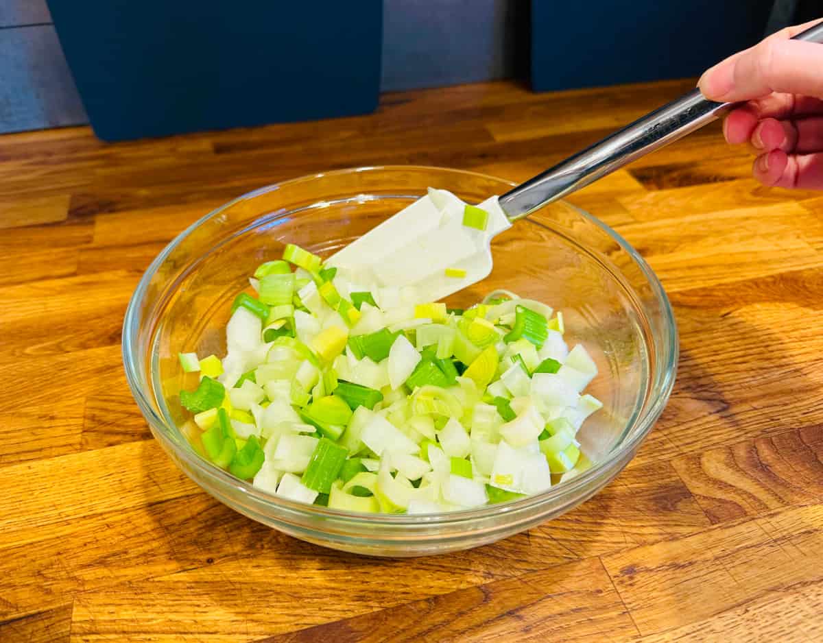 Chopped onion, celery, and leek being stirred with a silicone spatula in a glass bowl.