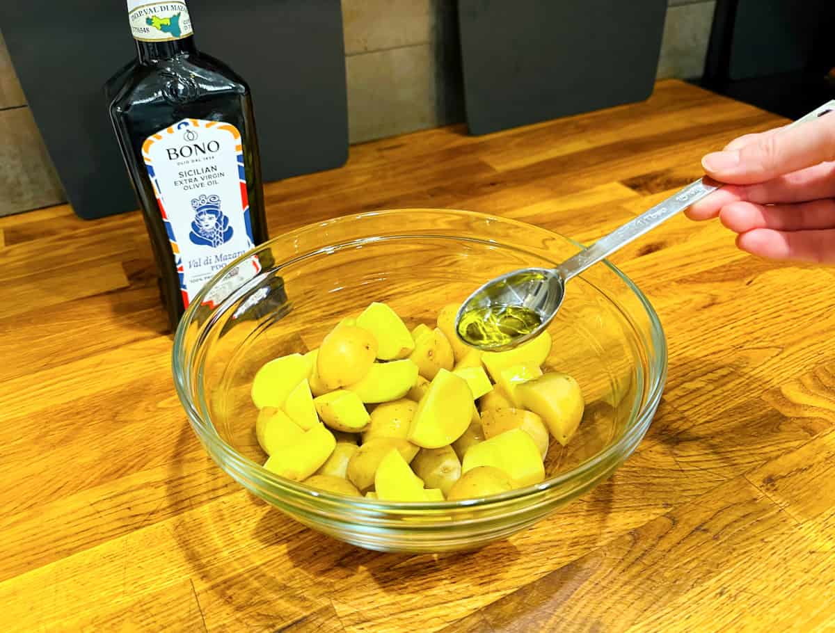 Olive oil being drizzled from a steel measuring spoon over quartered yellow potatoes in a glass bowl.