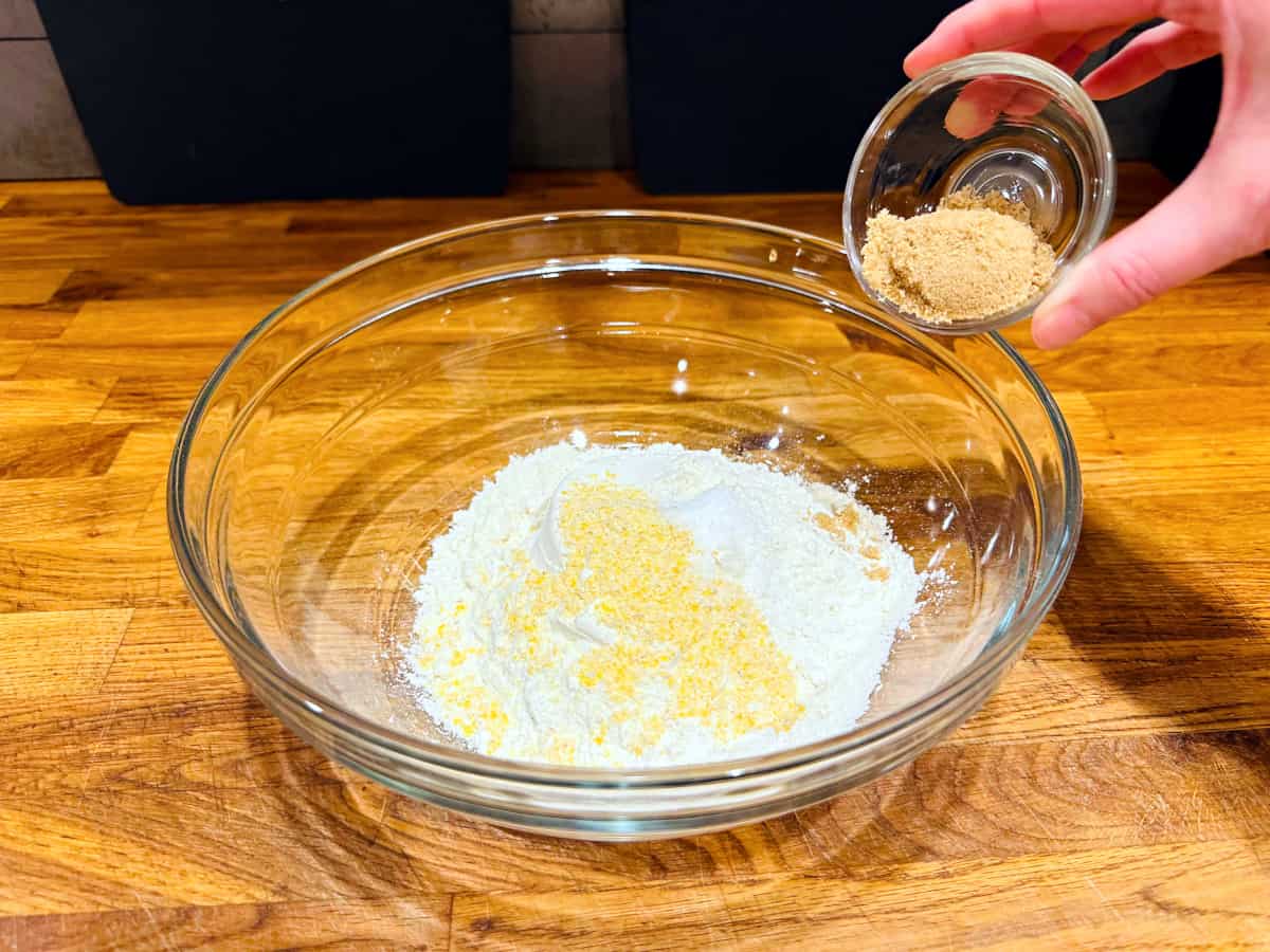 Light brown sugar being tipped from a tiny glass bowl into dry ingredients in a large glass bowl.