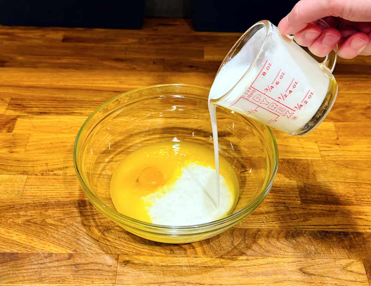 Buttermilk being poured from a glass measuring cup into mixture of melted butter and raw egg in a glass bowl.