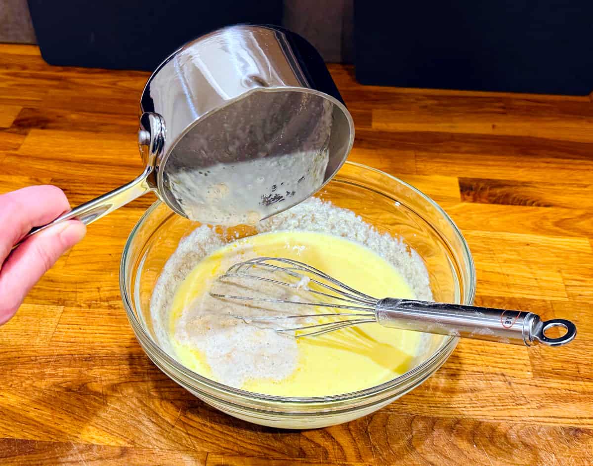 Milk and yeast mixture being poured from a small steel saucepan into a large bowl of yellow liquid with a steel whisk sitting in it.