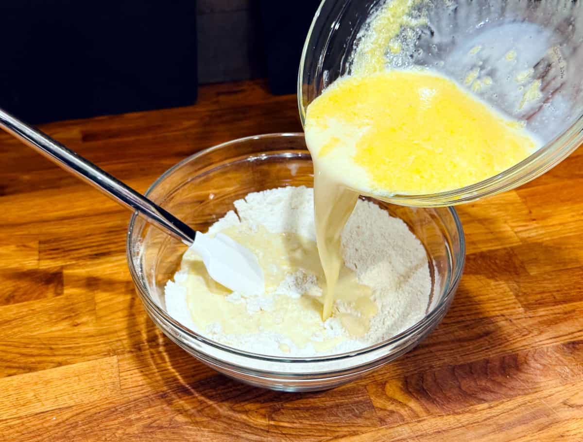 Pale yellow liquid being poured from a glass bowl into flour mixture in a large glass bowl.
