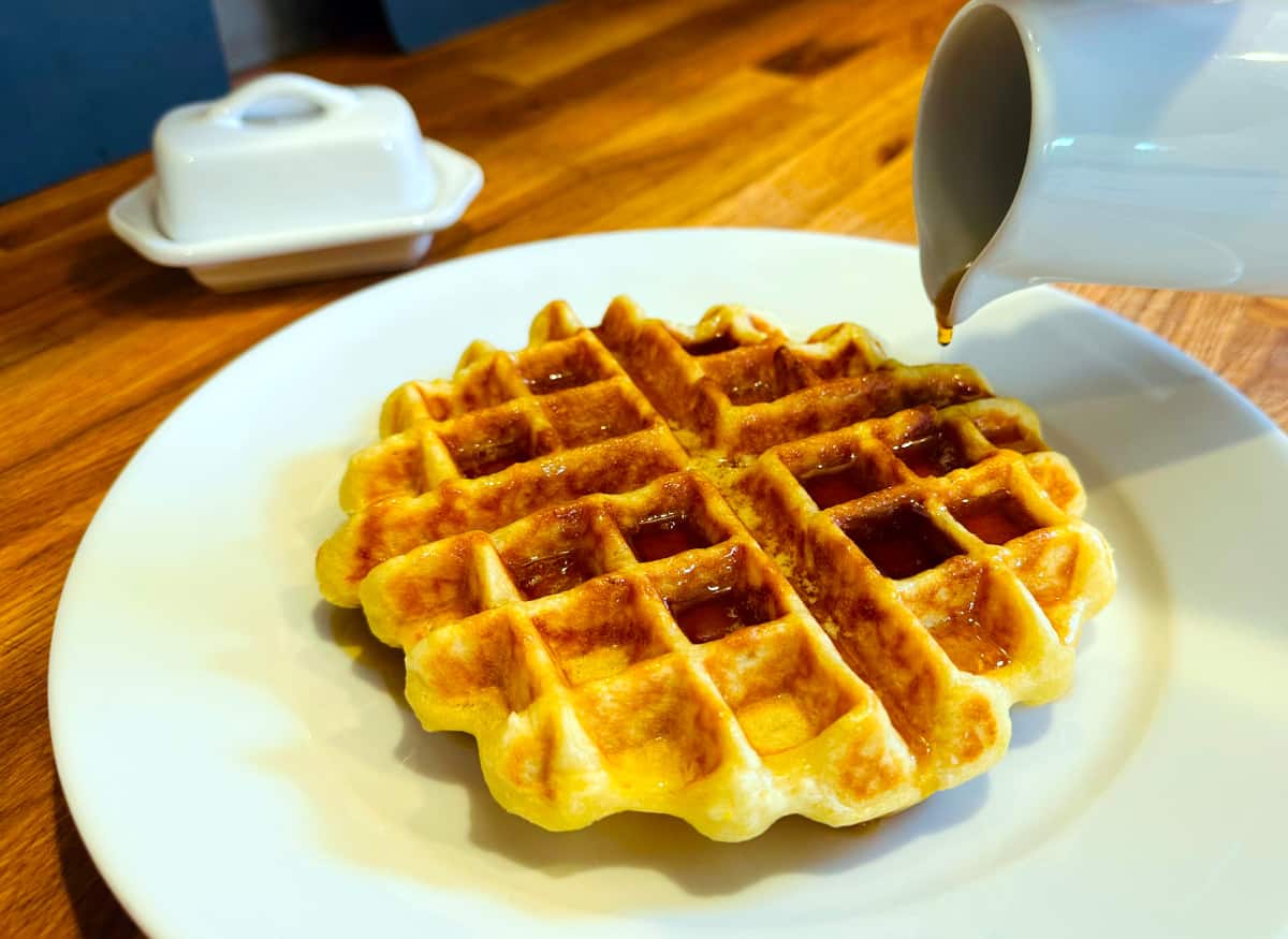 Maple syrup dripping from the tip of a white pitcher on to a homemade Belgian waffle covered with syrup on a white plate with a small butter dish in the background.